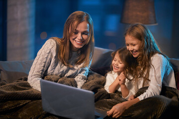 Fototapeta na wymiar Two little sisters and their mother sitting on couch under soft blanket and using modern laptop for watching cartoons. Happy family spending evening time with pleasure.