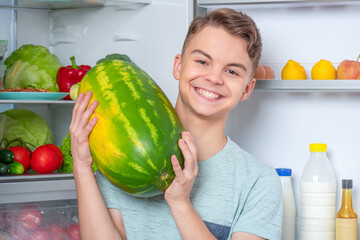 Fototapeta premium Handsome young teen boy holding big Watermelon while standing near open fridge in kitchen at home. Portrait of pretty child choosing food in refrigerator full of healthy products