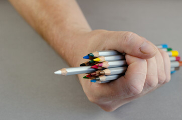 One hand holds a set of colored pencils on a gray background. A white pencil stands out. Multicolored watercolor pencils. Close-up. Selective focus.