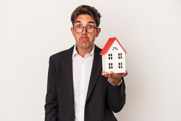 Fototapeta na wymiar Young mixed race business man holding a toy house isolated on white background shrugs shoulders and open eyes confused.
