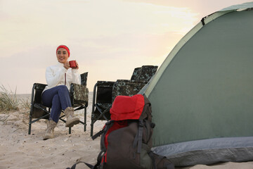 Young woman with cup of hot drink near camping tent on sandy beach