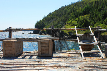 A wooden fish barrel with carrying frame on a pier. Atlantic Ocean and rugged coastline in background. Random Passage site, New Bonaventure, NL.