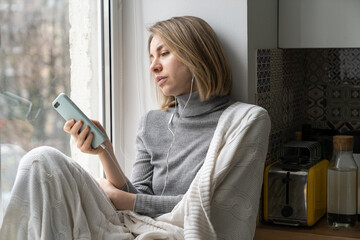 Uninterested woman wrapped in plaid sitting on windowsill at home, not in mood, using smartphone,...