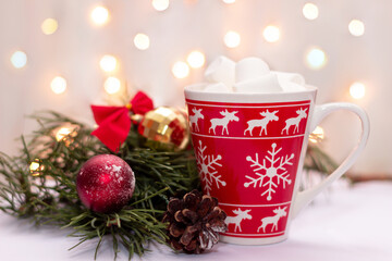A red mug with a deer, snowflakes pattern with beverage and marshmallow near a branch of a Christmas tree, toy balls and cone on a background of blurred bokeh lights of garland. New Year's decorations