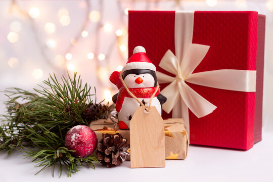 A toy Santa penguin with a wooden blank form for greeting text near a red gift box with a bow, a branch of a Christmas tree on a background of blurry bokeh lights of a garland. New Year's deorations 