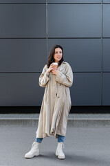 brunette woman in stylish autumn outfit standing with coffee to go and looking away