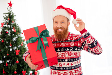 Photo of cheerful handsome male hold big red new year christmas present wear funky santa claus hat relaxing at home house