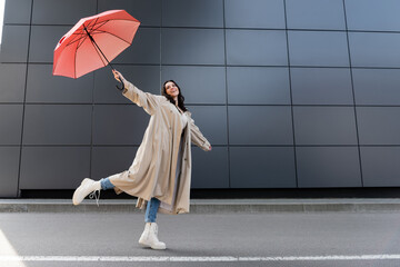 happy woman in beige coat and white boots posing with red umbrella in raised hand - obrazy, fototapety, plakaty
