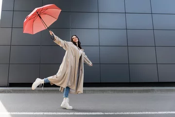 Fotobehang happy woman in beige coat and white boots posing with red umbrella in raised hand © LIGHTFIELD STUDIOS