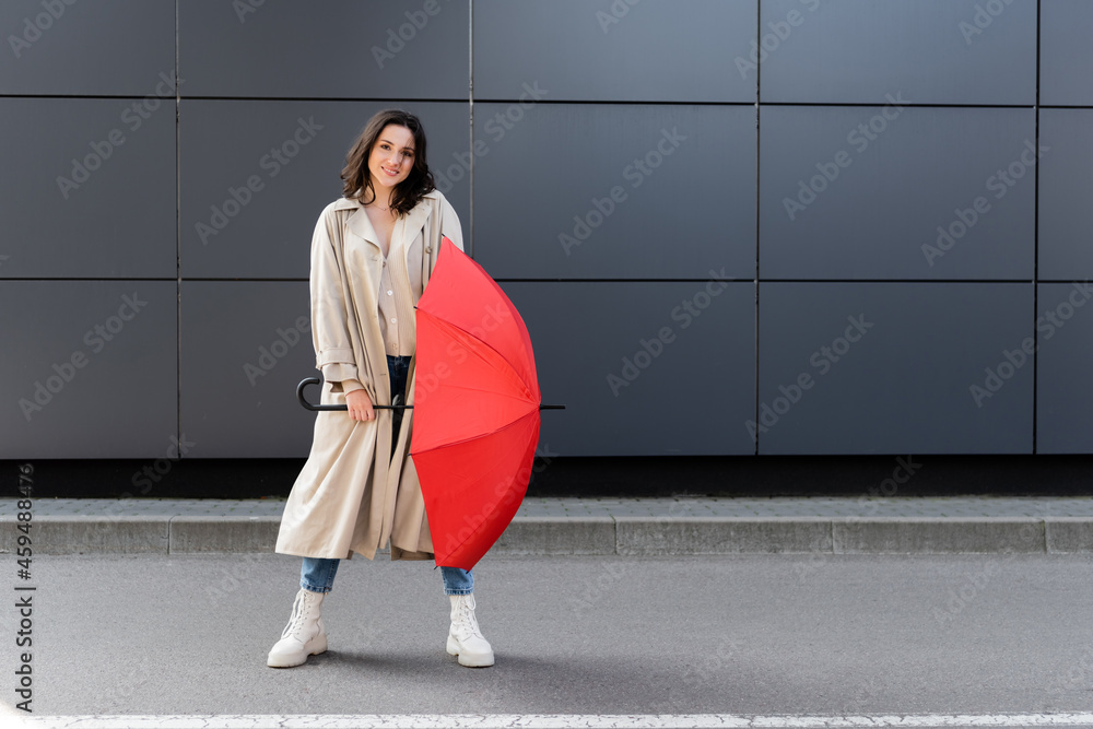 Wall mural trendy woman in long coat and white boots posing with red umbrella near grey wall