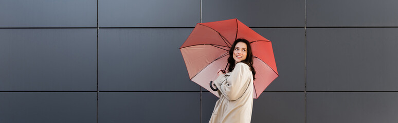 trendy woman with red umbrella looking back near grey wall, banner