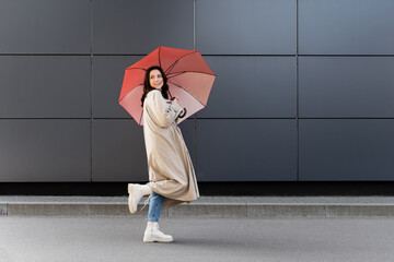 brunette woman in white boots and beige coat looking back under red umbrella