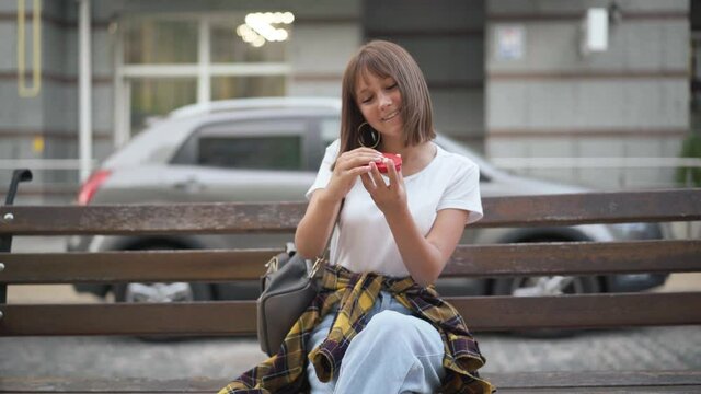 Portrait of charming slim beautiful teenage Caucasian girl with bob haircut admiring reflection in hand mirror smiling sitting on bench outdoors. Happy confident teenager on city street