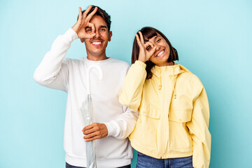 Fototapeta premium Young mixed race couple holding umbrella isolated on blue background excited keeping ok gesture on eye.