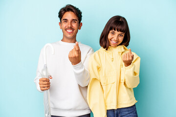 Young mixed race couple holding umbrella isolated on blue background pointing with finger at you as if inviting come closer.