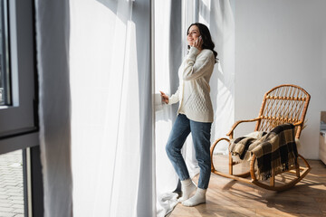full length view of woman in warm cardigan standing near window and talking on cellphone