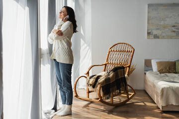 full length view of woman in jeans and white cardigan calling on smartphone near window and wicker chair - obrazy, fototapety, plakaty