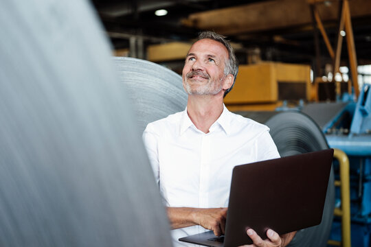 Smiling male inspector with laptop checking metal sheet roll in steel mill