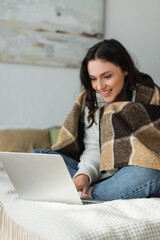 blurred woman smiling while using laptop on bed under checkered blanket