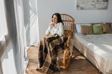 dreamy and smiling woman with cup of tea and book sitting in wicker chair under plaid blanket
