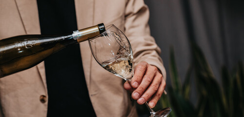Elegant waiter pours champagne from bottle into glass at restaurant. Sommelier tasting white...