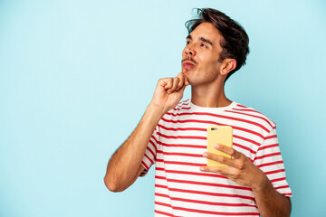 Young mixed race man holding mobile phone isolated on blue background looking sideways with doubtful and skeptical expression.