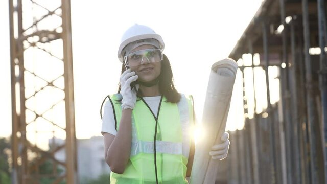  Indian female civil engineer talking on smartphone while holding blue prints at construction.Worker wearing hard hat at under construction building site outdoor.Asian woman wearing safety jacket.