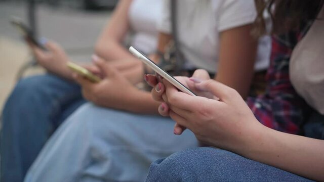 Close-up hands of teenage girls texting fast on smartphone sitting on city street outdoors. Unrecognizable device addicted absorbed Caucasian teenagers messaging online in social media