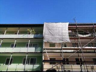 Baugerüst mit großer Plane als Schutz an der Fassade eines Wohnhaus mit Balkon vor blauem Himmel...