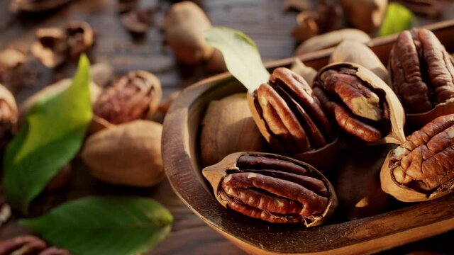 Pecans in a wooden bowl with pecan leaves and nut shells - close-up video, camera slow motion. The hand takes one of the pecan nut.