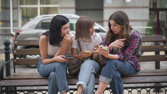 Positive joyful Caucasian teenage friends gossiping surfing social media on smartphones sitting on city street outdoors. Front view portrait of charming girls talking laughing sharing rumors