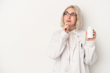 Young pharmacist woman holding pills isolated on white background looking sideways with doubtful...