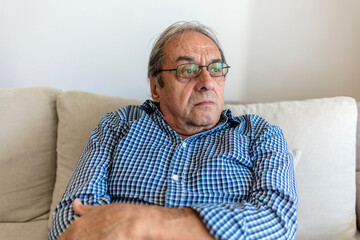 Shot of a senior man sitting by himself in a living room. Thoughtful elderly man sitting alone at home feeling lonely. Portrait of caucasian pensioner grandfather sitting at nursing home looking away.
