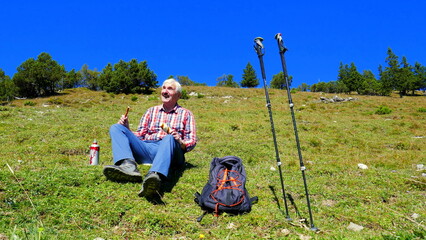 &auml;lterer Mann rastet fr&ouml;hlich auf sonniger Bergwiese mit Kiefern  neben Rucksack und Trekkingst&ouml;cken