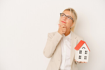 Young real estate agent woman holding a home model isolated on white background looking sideways...