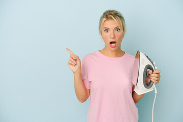 Young Russian woman holding iron isolated on blue background pointing to the side