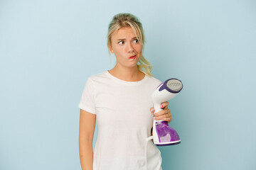 Young Russian woman holding vertical iron isolated on blue background confused, feels doubtful and unsure.