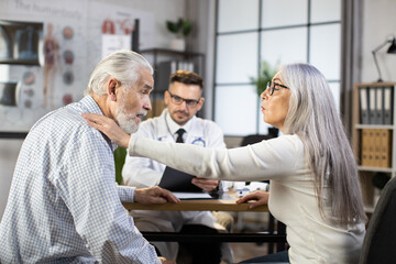 Obraz premium Caucasian aged woman supporting her husband visiting male doctor for regular checkup. Medical specialist in uniform and eyeglasses sitting at table and writing on clipboard.