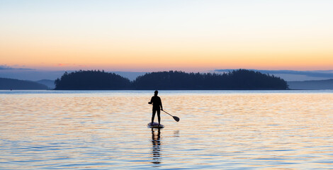 Adventurous Caucasian Adult Woman on a Stand Up Paddle Board is paddling on the West Coast of Pacific Ocean. Sunny Sunrise. Victoria, Vancouver Island, BC, Canada.