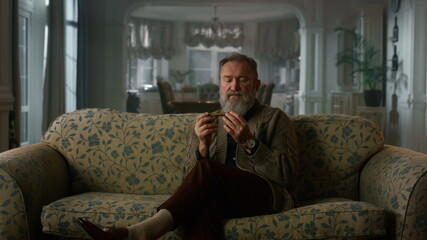 Relaxed senior gentleman smelling cuban cigar. Rich old man relaxing sofa