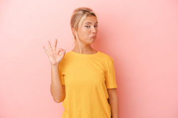 Obraz premium Young Russian woman isolated on pink background winks an eye and holds an okay gesture with hand.