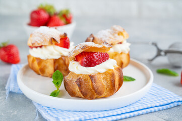 Delicious homemade small cakes profiterole choux pastry with custard, strawberry and icing powder on a gray concrete background. Copy space.