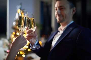 Smiling caucasian man in black suit holding glass of champagne and toasting with his wife while celebrating new year at home. Shiny christmas tree on blur background.