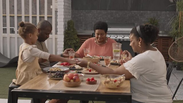 Medium of five Black family members of different generations sitting at table, all holding hands together and praying in silence before having dinner