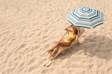 Woman resting in sunbed under striped beach umbrella at sandy coast, space for text