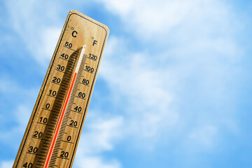 Wooden thermometer with red measuring liquid showing high temperature over 32 degrees Celsius on background of blue sky with clouds. Concept of heat wave, warm weather, global warming, climate change.