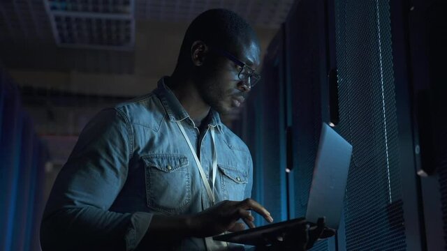 Young man working with laptop and checking equipment in dark server room spbas. Close-up view of American African male engineer holds device in his hand and does work, looks at racks with hardware and