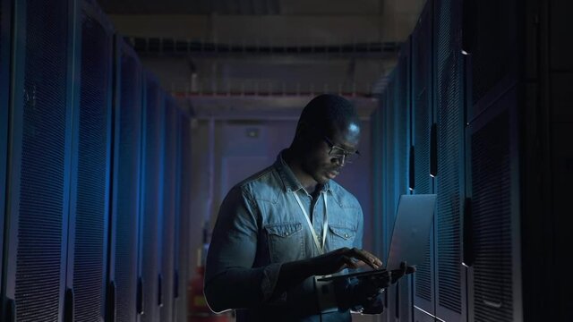 Male programmer working with laptop and supporting service while standing in data center spbas. Young African American man holds device in hand and looks at screen, inspects equipment or hardware rack