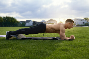 Muscular man with naked torso standing in plank position on yoga mat outdoors. Caucasian bodybuilder doing exercises for abs on grass of stadium. 