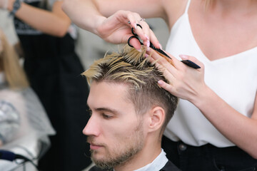 hairdresser cuts the hair of a young man with blond dyed hair with scissors in a beauty salon.